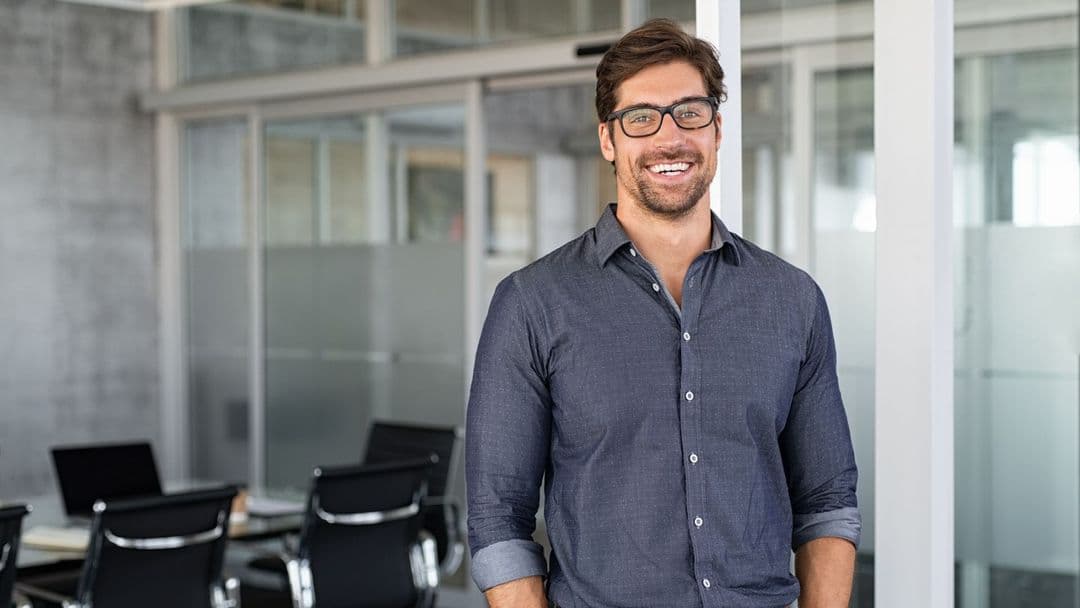 Ein Anwalt mit Brille und lockerem Hemd steht lächelnd in einem modernen Büro